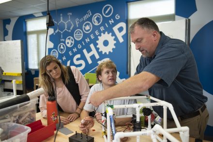 A professor works with a male and female student inside an engineering workship